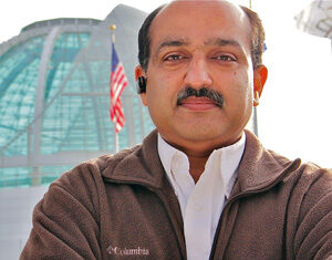 A man with a mustache stands with arms crossed, wearing a brown Columbia fleece jacket and a white shirt, in front of a glass building and a U.S. flag.