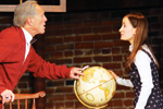 An older man and a young woman have a conversation indoors; the man gestures toward a globe while the woman holds it, with a brick wall background behind them.