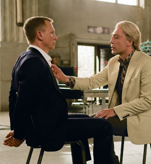 Two men sit facing each other in a dimly lit room. One man, in a dark suit, has his hands tied behind his back. The other, in a light-colored suit, leans forward, touching the first mans chest while speaking intensely.