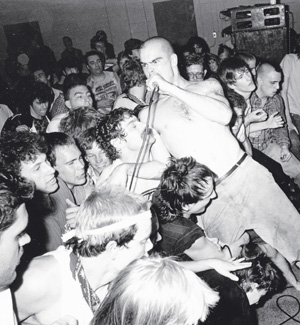 A black-and-white photo of a shirtless singer performing energetically in front of a packed, enthusiastic crowd at a small indoor concert or music venue. Microphone cables are visible amid the tightly packed audience.