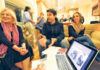 A group of people sit and talk in a cozy living room; three people smile and converse on a couch while others chat in the background. An open laptop with images on the screen is in the foreground.