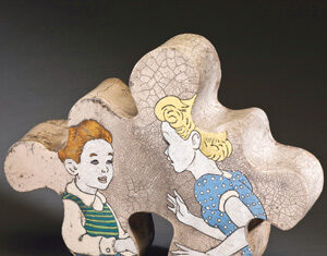 A ceramic sculpture shaped like an oversized jigsaw puzzle piece features painted cartoon-style images of a boy with red hair in a green striped shirt and a blonde girl in a blue polka-dot dress facing each other.