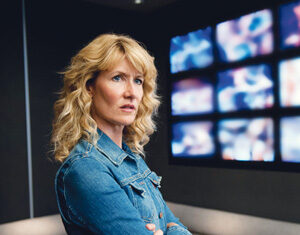 A woman with wavy blonde hair wearing a denim jacket stands with arms crossed in a dimly lit room, with multiple blurred screens displayed on the wall behind her.