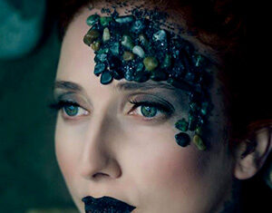 A woman with pale skin and green eyes has black lipstick and dark makeup. Her forehead and part of her cheek are decorated with small stones and textured black makeup, creating an artistic, nature-inspired look.
