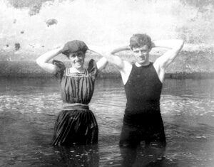 A black-and-white photo of a woman and a man standing waist-deep in water, both with their hands behind their heads, wearing old-fashioned bathing suits and smiling at the camera.
