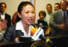 A woman speaks at a podium with microphones, addressing an audience. Several people stand behind her, some applauding. The setting appears to be an official or public event.