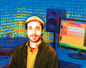 A man wearing a beige cap and dark jacket sits in a colorful, soundproof music studio with audio equipment, mixing consoles, and a computer monitor in the background.