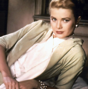 A woman with blonde hair pulled back, wearing a light green cardigan, white blouse, pearl earrings, and a pearl necklace, sits leaning slightly back with a calm expression.