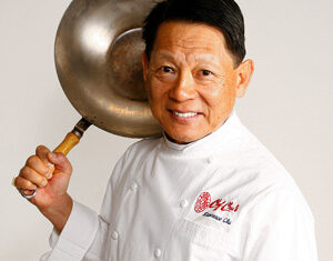 A smiling chef in a white coat holds a wok over his shoulder. The chef stands against a plain white background.