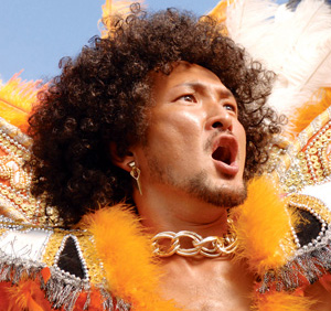 A person with curly hair and a beard wears dramatic makeup, a gold chain, feathered costume accents, and beaded details, looking upwards with their mouth open in a vibrant, expressive pose.