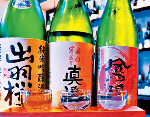 Three bottles of Japanese sake with colorful labels are displayed behind three filled sake glasses on a wooden tray, set on a bar with shelves of bottles in the background.