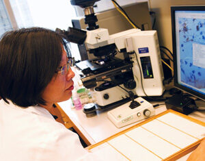 A scientist in a white lab coat examines data on a computer screen next to a microscope in a laboratory.