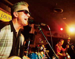 A musician wearing sunglasses sings into a microphone and plays an electric guitar on stage, with a drummer and upright bassist performing in the background under colorful stage lights.