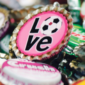 A close-up of a pink bottle cap with the word LOVE on it, where the letter O is replaced by a black and white soccer ball. Other colorful bottle caps are blurred in the background.