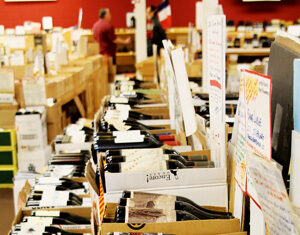 Cardboard boxes filled with wine bottles are arranged on display tables in a store with handwritten price tags and signs. A person stands in the background near shelves and a red wall.
