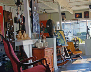 A sunlit art gallery interior with various paintings, sculptures, and decor items. A red rocking chair is in the foreground, with more furniture and artwork displayed throughout the space. Large windows let in natural light.