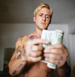 A shirtless man with blonde hair and visible tattoos intently counts a stack of dollar bills in a dimly lit room. The focus is on the money in his hands.