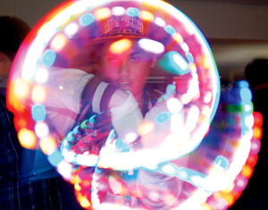 A person wearing a cap moves glowing lights in circular patterns, creating colorful, swirling light trails in a dark indoor setting.