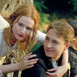 A young woman with long red hair leans affectionately on the shoulder of a young man with light brown hair. They both smile and appear to be sitting outdoors, near a tree, in a warm, sunlit setting.