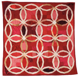 A handmade quilt featuring an interlocking ring pattern in shades of red, pink, and white, forming a geometric design on a square background.