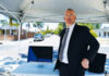A man in a suit stands under a canopy on a street, next to a table with two closed laptops. Houses, trees, and cars line the background on a sunny day.