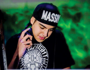 A young person wearing a cap labeled MASSIVE and headphones around their neck stands behind a laptop decorated with stickers, appearing to DJ in a vibrant, green-lit environment.