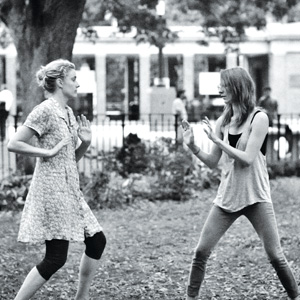 Two women stand facing each other in a park, both with their hands raised in defensive poses, as if practicing martial arts or playfully sparring. Trees and a fenced area are visible in the background.