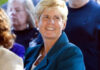 A smiling person with short blond hair wearing a teal blazer stands among a group of people, with others visible blurred in the background.