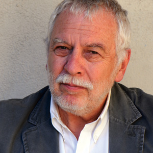 An older man with white hair and a beard, wearing a white shirt and a gray blazer, sits in front of a light-colored textured wall, looking at the camera with a slight smile.