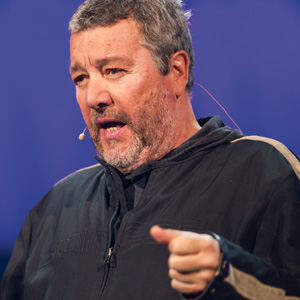 A man with gray hair and a beard speaks into a headset microphone, wearing a black jacket and gesturing with his hand against a blue background.