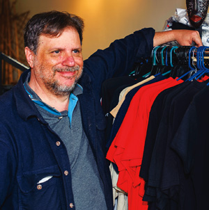A man with a beard and mustache smiles while standing next to a rack of assorted colorful shirts on hangers. He is wearing a blue jacket over a gray shirt.