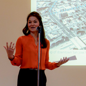 A woman in an orange blouse speaks into a microphone while holding papers, standing in front of a screen displaying a map of downtown San Jose.