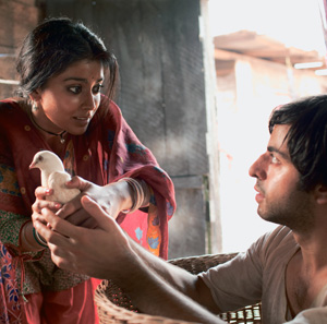 A woman holding a white dove looks surprised while showing it to a seated man, who looks up at her. They are indoors, near a doorway, with light streaming in.