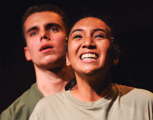 Two young adults on stage: a woman in front smiling widely and a man behind her looking slightly upward with a serious expression. Both wear casual shirts and are lit by stage lighting against a dark background.