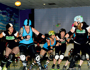 A group of women roller derby players in helmets and pads compete on an indoor rink, wearing uniforms with different colors and logos as they skate closely and interact physically.
