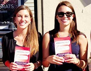 Two women standing side by side outdoors, smiling and holding red and white booklets. One is wearing sunglasses and a dark dress, the other is wearing a black jacket over a floral dress. Sunlight is shining on them.