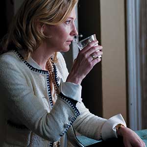 A woman with blonde hair in a white jacket with black trim sits by a window, holding a clear plastic cup near her mouth and looking thoughtfully outside.