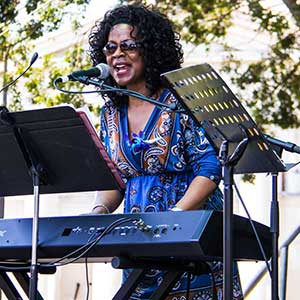 A woman wearing sunglasses and a patterned blue dress plays a keyboard and sings into a microphone on an outdoor stage, with trees in the background and music stands in front of her.