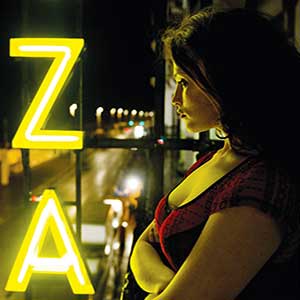 A woman with dark hair stands on a balcony at night, looking out over a street. Bright yellow neon letters Z and A are illuminated beside her. The background shows blurred lights of passing cars.