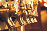 Close-up of several shiny brass beer taps in a row, with warm lighting and blurred colorful lights in the background.