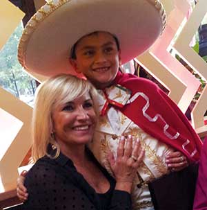 A smiling woman with blonde hair hugs a young boy dressed in a festive outfit with a large white sombrero and a red cape. They are indoors with decorative lighting in the background.