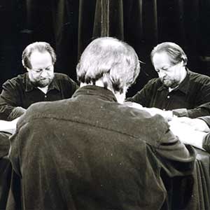 A man sits at a table with his back to the camera, looking down; his reflection appears in two mirrors, showing his face from different angles. The background is dark and the scene is in black and white.