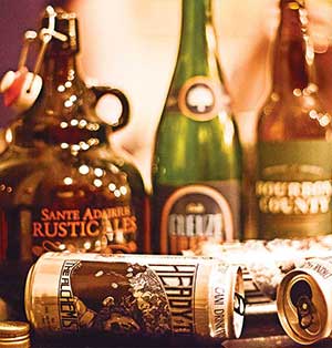 A variety of beer bottles and cans, including a jug labeled Sante Adairius Rustic Ales, sit on a table with blurred, warm lighting in the background.