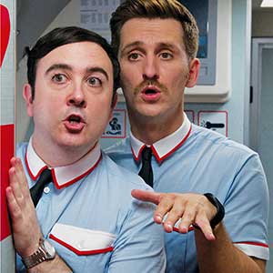 Two men in matching light blue uniforms with red and white trim stand close together, looking surprised or alarmed. One holds his hand up, the other gestures with both hands, as if reacting to something off camera.