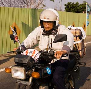 An older man wearing a white helmet and light jacket rides a motorcycle carrying bundles secured on the back, on a road next to a green fence.