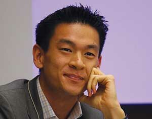 A man in a gray suit jacket and checkered shirt smiles while resting his chin on his hand during a panel or discussion, with a blurred purple and white background behind him.
