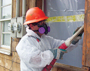 A construction worker in a hard hat, respirator, gloves, and protective suit uses equipment to remove material from a wooden house exterior near a window covered with plastic sheeting.