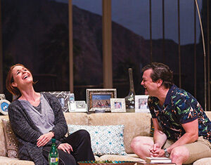 A woman and a man sit laughing together on a beige sofa in a living room decorated with framed photos and pillows. A glass bottle and books are on the wooden coffee table in front of them.