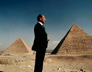 A man in a suit stands on sandy ground in front of two large pyramids under a clear blue sky, with the pyramids in the background.