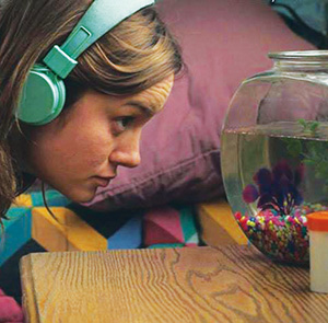 A woman wearing teal headphones closely observes a fishbowl on a wooden table, with colorful stones at the bottom, against a background of pillows and a patterned blanket.